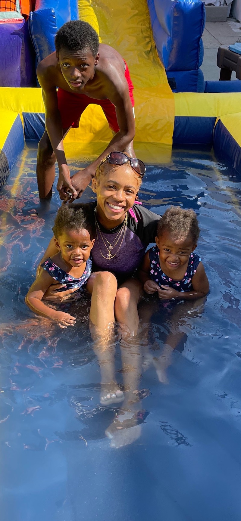 A teacher is with two little girls in the pool, and a boy in red shorts is smiling behind them after coming down the water slide.