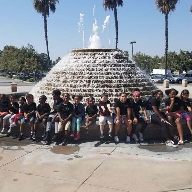 Kids from The Learning Box Child Enrichment Center sit in a row smiling for a picture in front of a fountain.