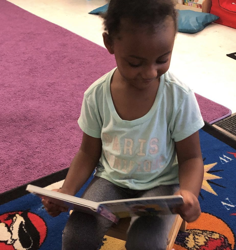 A little girl is sitting in the library reading a book at The Learning Box.