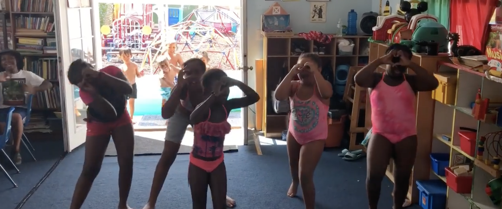 Five girls are making hearts with their hands while dancing, wearing their swimsuits after being in the pool.