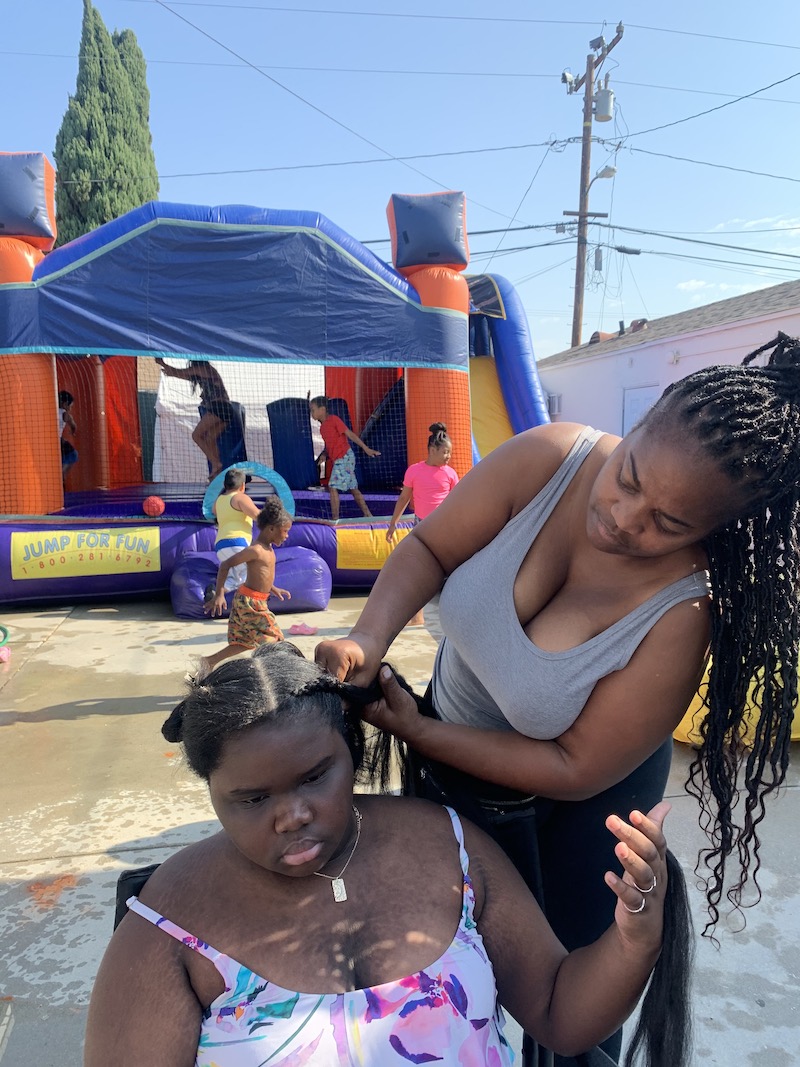Ms. Tee is braiding a girl's hair while children play in a bounce house in the background.