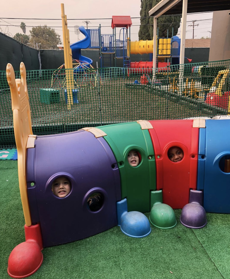 Three kids are smiling while looking out of the Climb-N-Crawl Caterpillar outdoor play structure peepholes on The Learning Box playground.