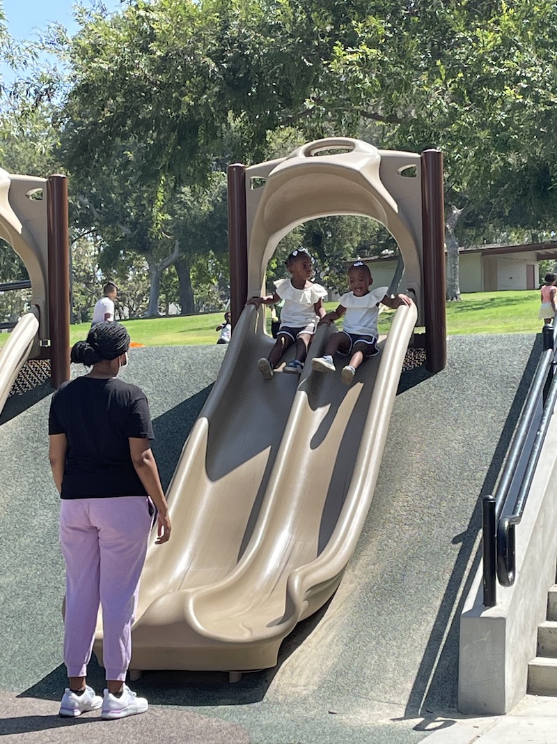 Two little girls are sliding down the slide with their teacher waiting to catch them.